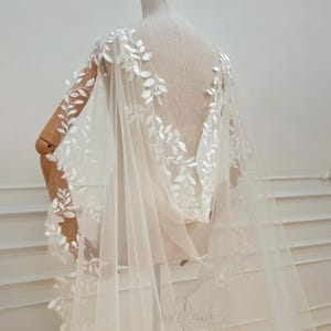 May include: A sheer, ivory-colored bridal cape with delicate white leaf embroidery. The cape is draped over a mannequin, showcasing the intricate floral design along the edges and back. The fabric is lightweight and flowing, ideal for weddings.