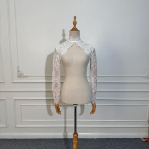 Op de afbeelding: Een witte kanten bolero met lange mouwen wordt getoond op een mannequin. De bolero heeft een hoge, gedetailleerde halslijn en ingewikkelde bloemen kanten patronen. De mannequin staat op een houten standaard, tegen een neutrale achtergrond.