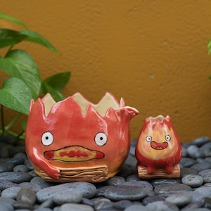 May include: Two ceramic planters, one large and one small, in the shape of a cartoon fire character. The large planter has a wide opening and a detailed face. The small planter is a miniature version. Both are red with white and brown accents.