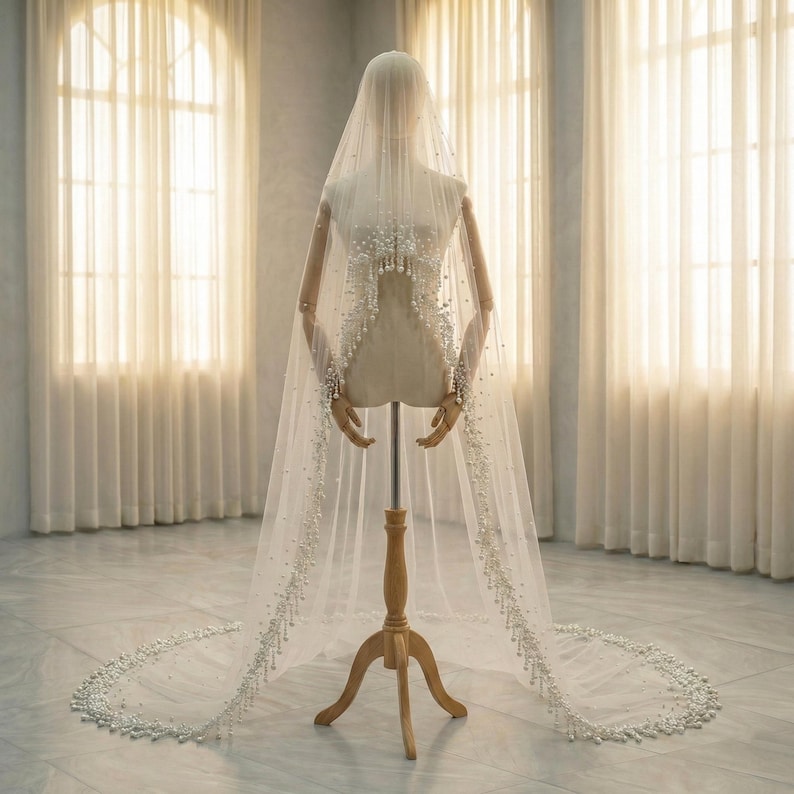 May include: A sheer white bridal veil with delicate pearl embellishments. The veil is draped over a mannequin, showcasing its flowing design. The pearls are clustered at the top and cascade down the edges, creating a beautiful, elegant look. The veil is long and trails on the floor.