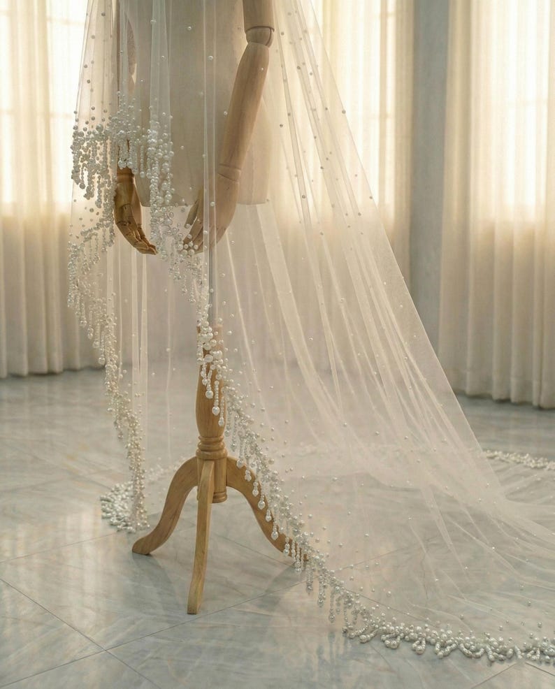 May include: A sheer white bridal veil adorned with scattered pearls and a beaded pearl trim. The veil is displayed on a wooden mannequin, showcasing its delicate fabric and elegant design. The veil's length extends to the floor.