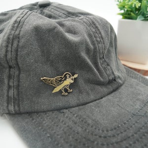 K&ouml;nnte beinhalten: Eine schwarze und goldene Emaille-Anstecknadel mit einer Kr&auml;he, die ein Schwert in ihrem Schnabel h&auml;lt. Die Anstecknadel ist an einer grauen Baseballkappe befestigt.
