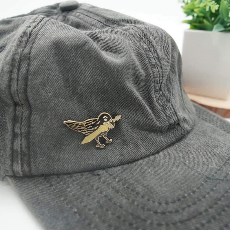 K&ouml;nnte beinhalten: Eine schwarze und goldene Emaille-Anstecknadel mit einer Kr&auml;he, die ein Schwert in ihrem Schnabel h&auml;lt. Die Anstecknadel ist an einer grauen Baseballkappe befestigt.