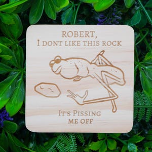 May include: A square wooden coaster with a frog illustration and the text "ROBERT, I DONT LIKE THIS ROCK, IT'S PISSING ME OFF". The frog is drawn in a cartoon style, with a rock next to it.