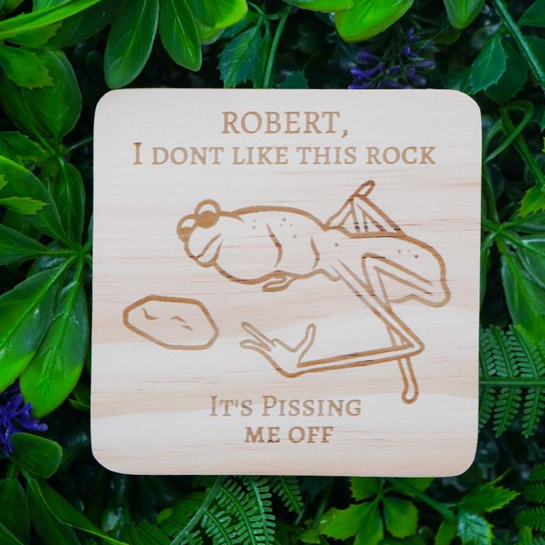 May include: A square wooden coaster with a frog illustration and the text "ROBERT, I DONT LIKE THIS ROCK, IT'S PISSING ME OFF". The frog is drawn in a cartoon style, with a rock next to it.