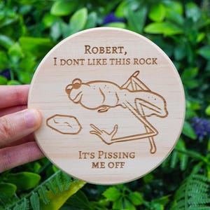 May include: A round wooden coaster with a frog illustration and the text "Robert, I DONT LIKE THIS ROCK. IT'S PISSING ME OFF." The coaster is light brown with a laser-etched design.