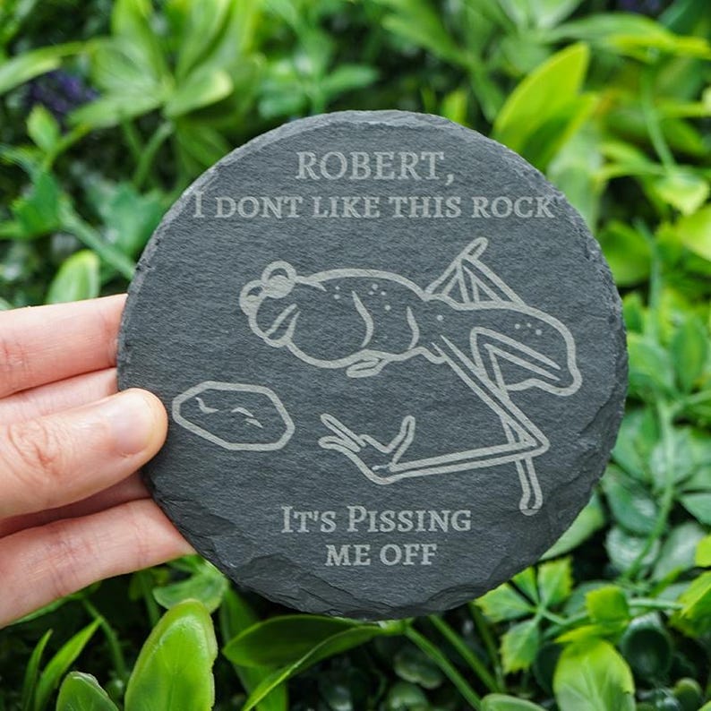 May include: A round, dark gray slate coaster with etched text and a frog illustration. The text reads "ROBERT, I DONT LIKE THIS ROCK" and "IT'S PISSING ME OFF". The frog illustration is in a humorous style.