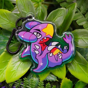 Peut inclure: Un porte-clés de personnage de dessin animé violet et jaune avec un fermoir noir. Le personnage a une marque en forme d'étoile et une bouche rouge. Le porte-clés est entouré de feuilles vertes.