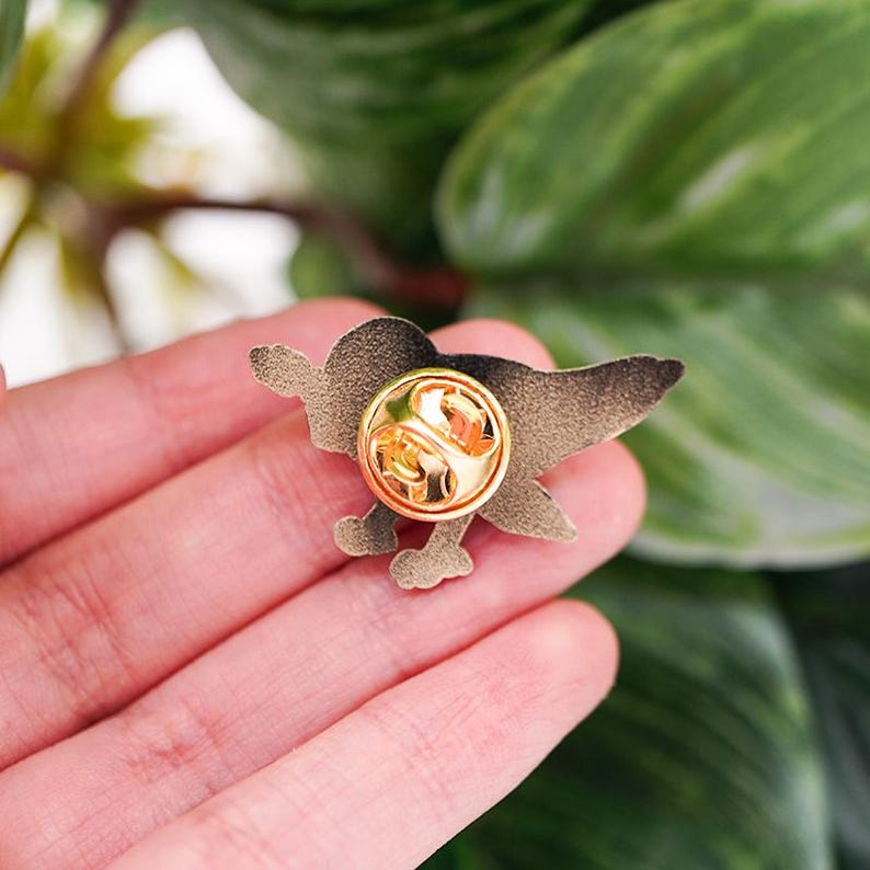 K&ouml;nnte beinhalten: Goldfarbene Emaille-Anstecknadel mit einem Cartoon-Vogel mit schwarzem Umriss. Die Nadel hat auf der R&uuml;ckseite einen Sicherheitsverschluss.
