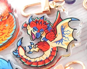 May include: A clear acrylic keychain featuring a colorful illustration of a dragon with red, yellow, and blue scales, a sharp beak, and wings. The charm is attached to a gold-toned metal lobster clasp hardware for easy attachment.