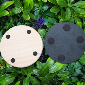 May include: Two round coasters. One is light wood with four black rubber feet. The other is dark gray with four black rubber feet. The coasters are displayed on a background of green and purple foliage.