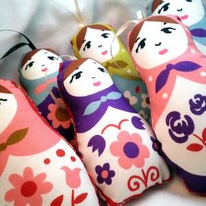 May include: A collection of handmade fabric dolls in the style of Matryoshka nesting dolls. Each doll features a painted face, brown hair, and a colorful floral pattern. The dolls are in shades of pink, purple, blue, and green, with white and red accents.