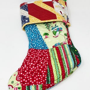 May include: A patchwork Christmas stocking with a green loop for hanging. The stocking is made of various fabrics with red, green, blue, yellow, and white patterns. The stocking is quilted and has a festive design.