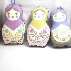 May include: Five decorative fabric dolls in various colors, including teal, purple, and yellow. Each doll features a painted face and a floral apron design. They have blue hanging loops.
