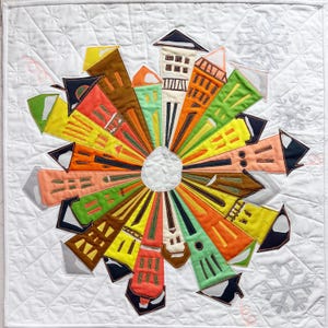 May include: A colorful, handmade quilt featuring a circular design of stylized buildings radiating from a central point. The buildings are various colors, including orange, green, and brown, set against a white background with snowflake and diamond quilting.