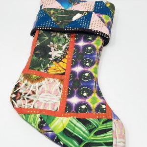 May include: A colorful patchwork Christmas stocking with a green loop handle. The stocking is made of different fabrics with floral, butterfly, and geometric patterns. The stocking is quilted and has a red border.
