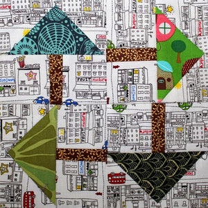 Op de afbeelding: Een patchwork quiltblok met een stadsthema. Het blok is gemaakt van verschillende stoffen met verschillende patronen, waaronder een groen-wit bloemenpatroon, een bruin-gouden bloemenpatroon, een turquoise-wit geometrisch patroon en een groen-wit bladerenpatroon. Het blok heeft ook een witte achtergrond met zwarte contouren van gebouwen en andere stedelijke elementen.