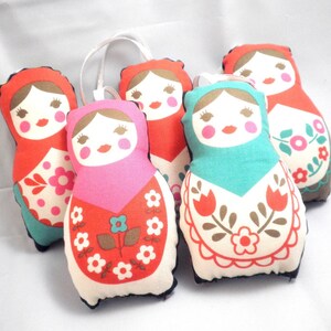 May include: A group of five fabric Matryoshka dolls. Each doll features a white face with rosy cheeks, a red or pink head covering, and a patterned apron. The aprons display floral designs in red, pink, and teal. A white loop is attached to the top.