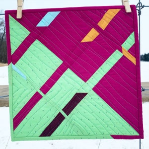 May include: A small quilt with geometric shapes in shades of green, pink, orange, and purple. The quilt is made of fabric squares sewn together to create a patchwork design.