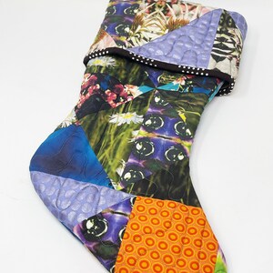 May include: A colorful patchwork Christmas stocking with a green loop at the top. The stocking is made of various fabrics with floral, geometric, and abstract patterns in shades of blue, purple, green, orange, and white.