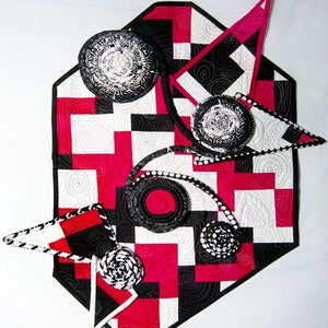 May include: A black and white quilted table runner with a geometric pattern and pink accents. The runner is decorated with several black and white fabric rosettes.