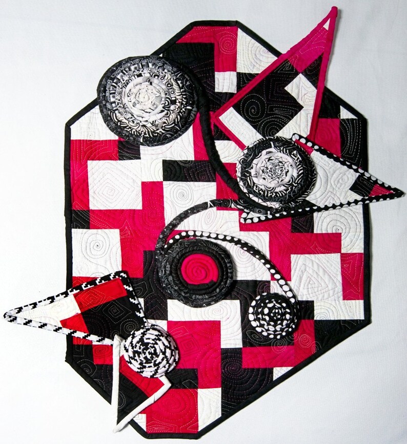 May include: A black and white quilted table runner with a geometric pattern and pink accents. The runner is decorated with several black and white fabric rosettes.