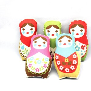 May include: Five colorful fabric Matryoshka dolls. Each doll has a painted face with rosy cheeks and a red mouth. The dolls are in shades of red, green, pink, blue, and yellow, with floral apron designs.