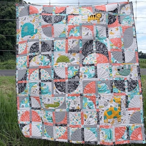 May include: A quilted blanket featuring a geometric pattern of semicircles and squares in black, white, coral, and teal. The fabric displays various animal illustrations including a yellow giraffe, a teal elephant, a green turtle, and a sloth.