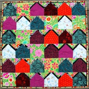 Puede incluir: Una colcha colorida con un patrón de casas. Las casas están hechas de diferentes telas y colores, incluyendo rojo, verde, azul, morado y blanco. La colcha está bordeada con un ribete marrón y blanco.