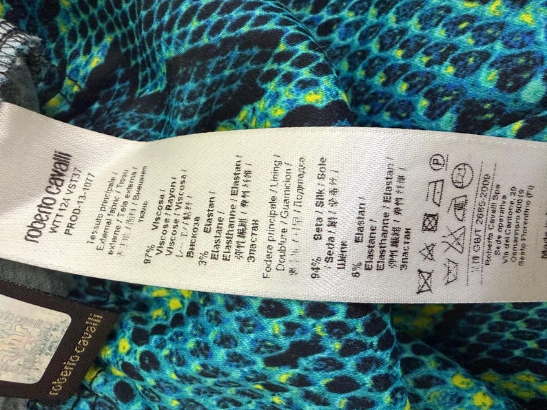 K&ouml;nnte beinhalten: Ein Roberto Cavalli Kleidungsetikett mit Stoffzusammensetzungsdetails, darunter 97% Viskose und 3% Elastan f&uuml;r das &Auml;u&szlig;ere sowie 94% Seide und 6% Elastan f&uuml;r das Futter, vor einem lebendigen blau-schwarz-gelben Schlangenmuster.