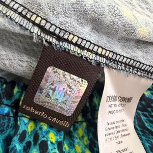 K&ouml;nnte beinhalten: Ein Roberto Cavalli Kleidungsetikett mit einem holografischen Echtheitsaufkleber mit dem Markenlogo. Das Etikett ist an einem Stoff mit blauem, schwarzem und gelbem Tiermuster befestigt. Ein wei&szlig;es Pflegeetikett listet 97% Viskose und den Code WTT124 VST37.