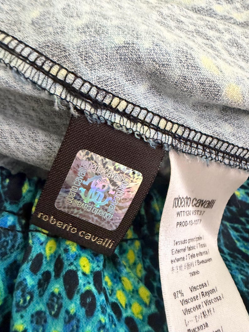 K&ouml;nnte beinhalten: Ein Roberto Cavalli Kleidungsetikett mit einem holografischen Echtheitsaufkleber mit dem Markenlogo. Das Etikett ist an einem Stoff mit blauem, schwarzem und gelbem Tiermuster befestigt. Ein wei&szlig;es Pflegeetikett listet 97% Viskose und den Code WTT124 VST37.