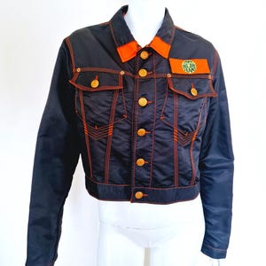 May include: A black denim jacket with orange stitching and a small green and white logo patch on the left chest. The jacket has a button closure and two front pockets.