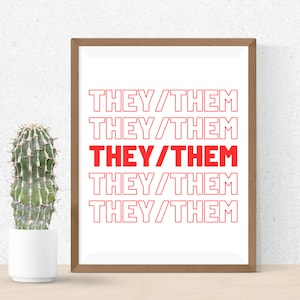 May include: A framed print with the text "THEY/THEM" in red, repeated five times. The print is in a light brown frame and is displayed on a wooden surface. A cactus in a white pot is to the left of the frame.
