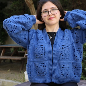 Könnte beinhalten: Eine blaue gehäkelte Strickjacke mit quadratischem Muster und V-Ausschnitt. Die Strickjacke hat lange Ärmel und ist vorne offen.