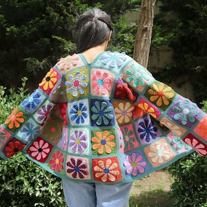 Könnte beinhalten: Eine bunte gehäkelte Strickjacke mit Blumenmuster. Die Strickjacke besteht aus Quadraten mit verschiedenen farbigen Blumen in jedem Quadrat. Die Strickjacke ist hellblau.