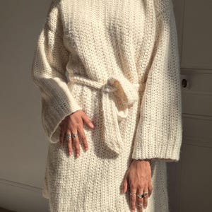 Peut inclure: Une robe pull en crochet de couleur crème avec des manches longues et une ceinture à nouer.