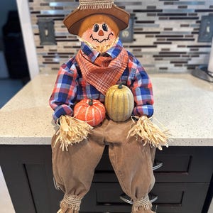 May include: A scarecrow figurine wearing a brown hat with burlap trim, a blue and red plaid shirt, an orange plaid neckerchief, and brown pants. The figure has straw-like hands and feet, and holds a small orange pumpkin and a tan pumpkin.
