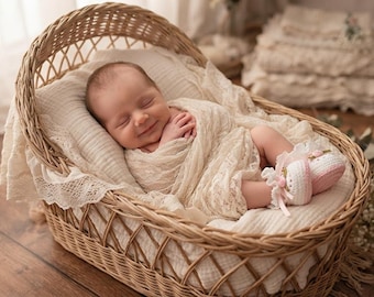 May include: A newborn infant sleeping in a woven wicker basket, wrapped in a cream-coloured lace swaddle. The infant wears small, white and pink crocheted booties with lace trim. The basket is lined with a soft, textured cream fabric and a pillow.