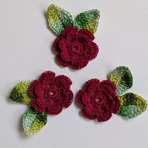 May include: Three handmade crochet flower appliqués, each featuring a deep burgundy red blossom with a central hole and two variegated green leaves. The leaves display a gradient of light to dark green tones, crafted with visible yarn stitches.