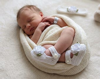 May include: A set of white crocheted baby booties featuring a satin rose and a pearl button closure, accompanied by a matching white crocheted headband with a central white rose and green leaf accent, all crafted from soft yarn.