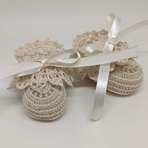 Puede incluir: Un par de botitas de bebé de ganchillo con delicados detalles de encaje y lazos de cinta de satén. Las botitas son de color crema y tienen un aspecto suave y femenino.