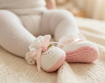 May include: A pair of white and light pink crocheted baby booties featuring a delicate white lace trim around the ankle, a soft pink satin ribbon bow, and a small decorative pink rose accent centred on the front of each shoe.