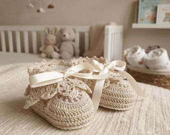 May include: A pair of beige crochet baby booties featuring a delicate scalloped edge around the ankle opening and a smooth cream-coloured satin ribbon tied in a bow across the top of each shoe, showcasing intricate yarn stitching throughout.