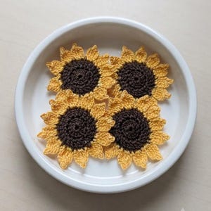 May include: Four crocheted sunflower appliqués arranged in a white ceramic bowl. Each sunflower features a dark brown circular centre surrounded by bright yellow petals, crafted with detailed yarn stitching to mimic the appearance of a flower.