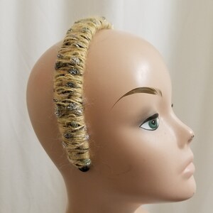 May include: A beige headband with silver and green embellishments. The headband is made of yarn and is wrapped around a mannequin's head.