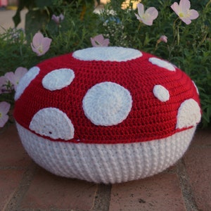 May include: A handmade crochet mushroom pillow. The top is red with white polka dots, and the base is white. The pillow is round and sits on a brick surface, with greenery and pink flowers in the background.