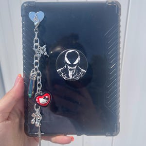 May include: A black tablet case featuring a circular Venom character pop socket and a silver-toned metal chain charm. The charm includes a heart-shaped clasp, a butterfly, a prismatic crystal, a Hello Kitty heart, cherries, and a sword pendant.