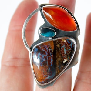 May include: A unique silver ring featuring a large, multi-colored gemstone with shades of brown, blue, and orange. The ring also has a vibrant orange stone and a small teal stone. The ring's design is abstract and modern.