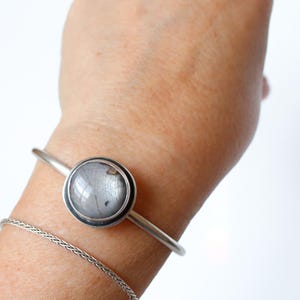 May include: A silver-toned bracelet with a large, round, gray stone set in a bezel. A delicate chain bracelet is also visible. The jewelry is displayed on a person's wrist against a white background. The stone appears to have internal patterns.